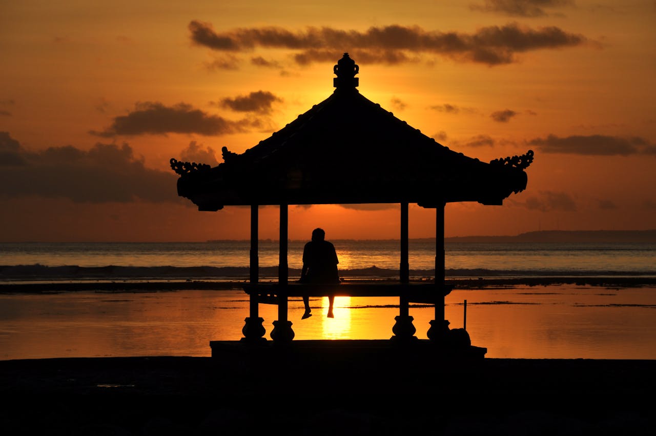 A serene silhouette of a man enjoying a stunning Bali sunset from a gazebo by the sea.
